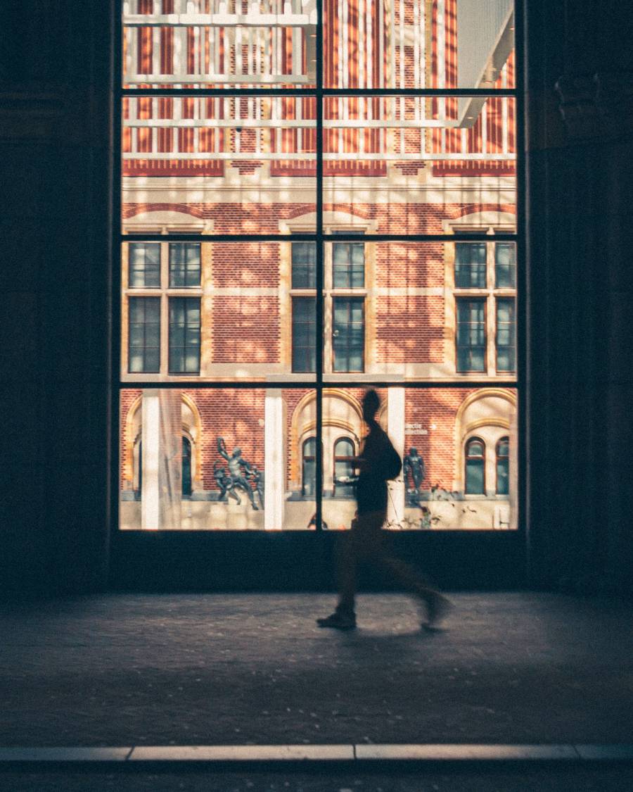 Silhouet van iemand die langs een hoog raam loopt, met daarachter de rode bakstenen gevel van het Rijksmuseum