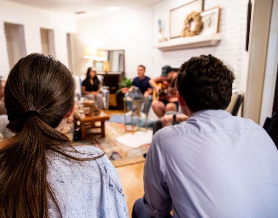 Een bijeenkomst in een huiskamer: twee mensen, van achteren gezien, luisteren naar iemand die gitaar speelt